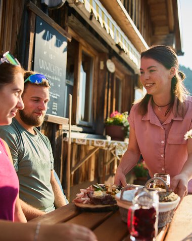 Drei Menschen genießen Sommer im Außenbereich einer Berghütte im Kleinwalsertal, während sie Mahlzeiten und Getränke auf einem Holztablett teilen. | © Kleinwalsertal Tourismus eGen • Fotograf: Oliver Farys