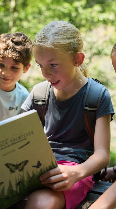 Drei Kinder sitzen im Wald und lesen gemeinsam ein Buch über Naturforschung, umgeben von grünen Bäumen an einem sonnigen Tag. | © Kleinwalsertal Tourismus eGen • Fotograf: Oliver Farys