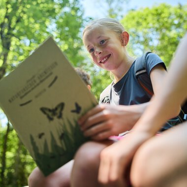 Ein lachendes Mädchen liest eine Naturbroschüre in einem gelben, grünen Wald im Sommer 2024. | © Kleinwalsertal Tourismus eGen • Fotograf: Oliver Farys
