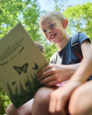 Ein lachendes Mädchen liest eine Naturbroschüre in einem gelben, grünen Wald im Sommer 2024. | © Kleinwalsertal Tourismus eGen • Fotograf: Oliver Farys