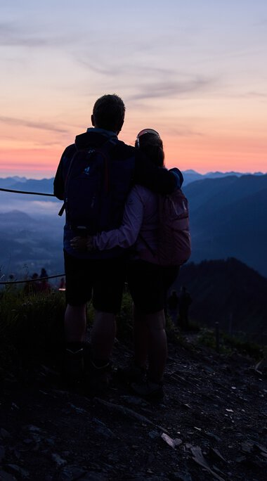 Ein Paar steht beim Sonnenaufgang in den Bergen und schaut in die Weite des Tals, umgeben von einer ruhigen Landschaft bei Sonnenaufgang. | © Kleinwalsertal Tourismus eGen • Fotograf: Oliver Farys