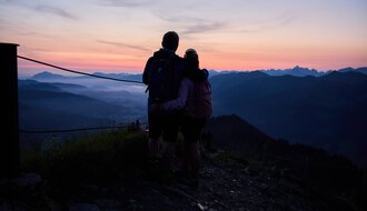 Ein Paar steht beim Sonnenaufgang in den Bergen und schaut in die Weite des Tals, umgeben von einer ruhigen Landschaft bei Sonnenaufgang. | © Kleinwalsertal Tourismus eGen • Fotograf: Oliver Farys
