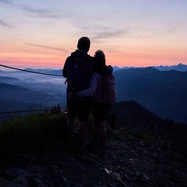 Ein Paar steht beim Sonnenaufgang in den Bergen und schaut in die Weite des Tals, umgeben von einer ruhigen Landschaft bei Sonnenaufgang. | © Kleinwalsertal Tourismus eGen • Fotograf: Oliver Farys