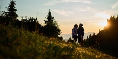 Zwei Menschen stehen beim Sonnenaufgang in einer Landschaft mit Bäumen und Hügeln und genießen die Natur im Kleinwalsertal. | © Kleinwalsertal Tourismus eGen • Fotograf: Oliver Farys