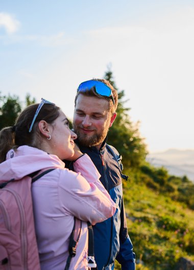 Ein Paar beim Wandern im Sonnenaufgang im Kleinwalsertal, umgeben von grünen Bergen und Bäumen, lachend und sich gegenseitig zugewandt. | © Kleinwalsertal Tourismus eGen • Fotograf: Oliver Farys