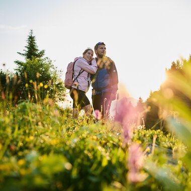 Ein Paar wandert in einem blühenden Feld bei Sonnenaufgang im Kleinwalsertal, umgeben von Natur und Bäumen. | © Kleinwalsertal Tourismus eGen • Fotograf: Oliver Farys