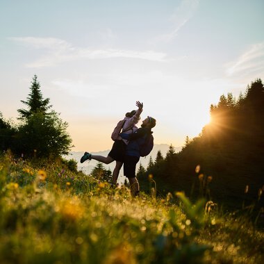 A couple lifts each other up on a green mountain meadow at sunrise in Kleinwalsertal, surrounded by trees, with sunlight in the background. | © Kleinwalsertal Tourismus eGen • Photographer: Oliver Farys