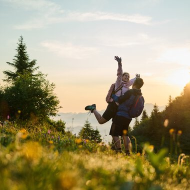 A couple hiking at sunrise in Kleinwalsertal, surrounded by nature and green fields, with the sun rising in the background. | © Kleinwalsertal Tourismus eGen • Photographer: Oliver Farys