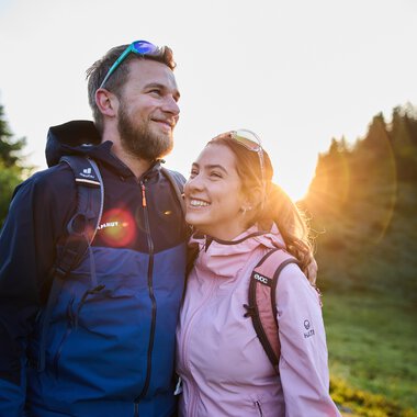 Ein lachendes Paar beim Wandern im Sommer, Sonnenaufgang im Hintergrund in einem Waldgebiet. | © Kleinwalsertal Tourismus eGen • Fotograf: Oliver Farys