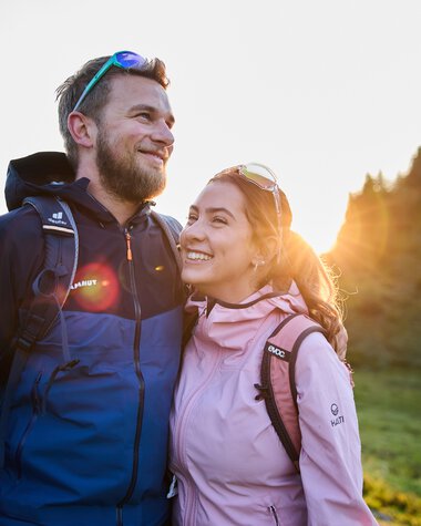 A smiling couple hiking during summer sunrise in a forested area. | © Kleinwalsertal Tourismus eGen • Photographer: Oliver Farys