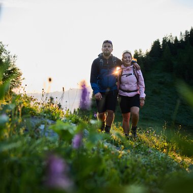 A couple hiking during sunrise in Kleinwalsertal, surrounded by flowers and green mountains. | © Kleinwalsertal Tourismus eGen • Photographer: Oliver Farys