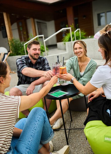 Four young adults sitting outdoors in summer, clinking drinks and laughing, enjoying a relaxed time outside their house. | © Kleinwalsertal Tourismus eGen • Photographer: Oliver Farys