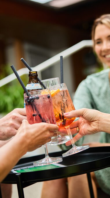 Drei Personen stoßen bei einem Sommer-Get-together mit bunten Drinks in Gläsern an, im Hintergrund eine lachende Frau im Café. | © Kleinwalsertal Tourismus eGen • Fotograf: Oliver Farys