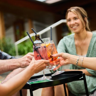 Drei Personen stoßen bei einem Sommer-Get-together mit bunten Drinks in Gläsern an, im Hintergrund eine lachende Frau im Café. | © Kleinwalsertal Tourismus eGen • Fotograf: Oliver Farys