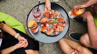 Drei Menschen sitzen um einen kleinen schwarzen Tisch im Sommer, trinken Getränke mit Zitronenscheiben und teilen eine Platte mit verschiedenen warmen Pizzastücken. | © Kleinwalsertal Tourismus eGen • Fotograf: Oliver Farys