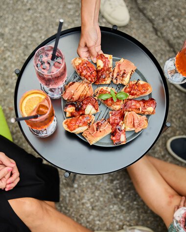 Drei Menschen sitzen um einen kleinen schwarzen Tisch im Sommer, trinken Getränke mit Zitronenscheiben und teilen eine Platte mit verschiedenen warmen Pizzastücken. | © Kleinwalsertal Tourismus eGen • Fotograf: Oliver Farys
