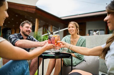 Vier Freunde sitzen im Sommergarten, lachen und stoßen mit Cocktails an, genießen eine entspannte Zeit im Kleinwalsertal. | © Kleinwalsertal Tourismus eGen • Fotograf: Oliver Farys
