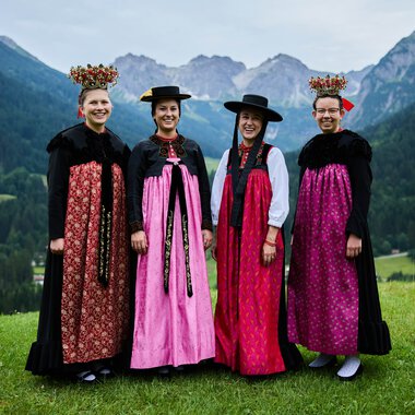 Four women in traditional costumes standing on a green meadow with mountains and forests in the background. | © Kleinwalsertal Tourismus eGen • Photographer: Oliver Farys