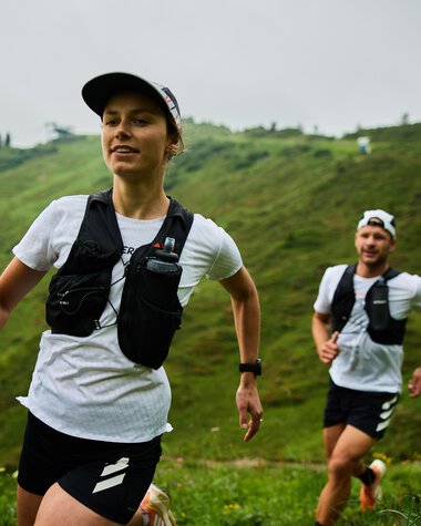Zwei Trailrunner, eine Frau und ein Mann, laufen auf einem grünen Hügel in Kleinwalsertal bei sommerlichem Wetter. Beide tragen Laufkleidung und Tragegurte. | © Kleinwalsertal Tourismus eGen • Fotograf: Oliver Farys