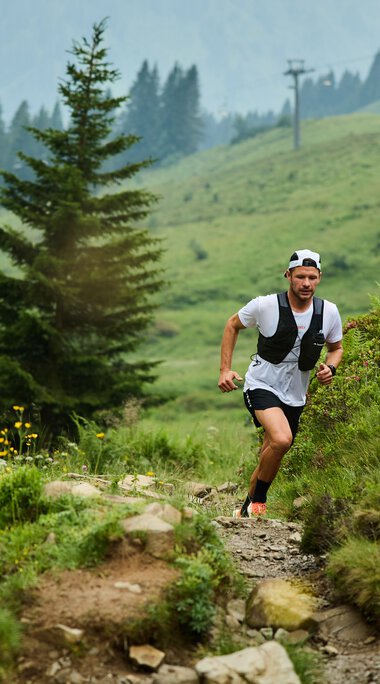 Zwei Trailrunner, ein Mann und eine Frau, laufen auf einem schmalen, felsigen Pfad in einer grünen Berglandschaft mit Bäumen und Blumen, im Hintergrund Berge und Wolken. | © Kleinwalsertal Tourismus eGen • Fotograf: Oliver Farys