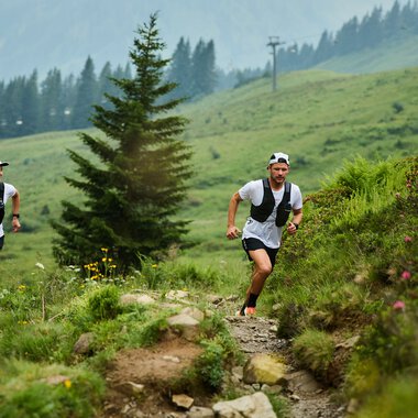Zwei Trailrunner, ein Mann und eine Frau, laufen auf einem schmalen, felsigen Pfad in einer grünen Berglandschaft mit Bäumen und Blumen, im Hintergrund Berge und Wolken. | © Kleinwalsertal Tourismus eGen • Fotograf: Oliver Farys