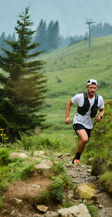 Zwei Trailrunner, ein Mann und eine Frau, laufen auf einem schmalen, felsigen Pfad in einer grünen Berglandschaft mit Bäumen und Blumen, im Hintergrund Berge und Wolken. | © Kleinwalsertal Tourismus eGen • Fotograf: Oliver Farys