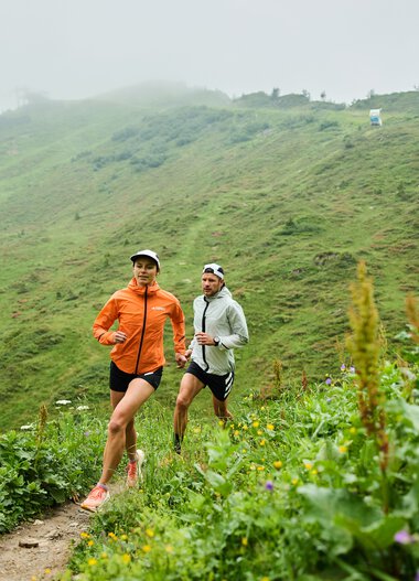 Zwei Läufer in Sportkleidung laufen auf einer grünen, hügeligen Trail in den Bergen des Kleinwalsertals, umgeben von Pflanzen und Blumen. | © Kleinwalsertal Tourismus eGen • Fotograf: Oliver Farys