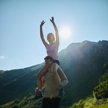 Ein Mann trägt ein Mädchen auf den Schultern, während sie bei sonnigem Wetter in einer Berglandschaft spazieren gehen, mit Bergen im Hintergrund. | © Kleinwalsertal Tourismus eGen • Fotograf: Oliver Farys