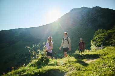 Drei Wanderer auf einem Weg im Kleinwalsertal bei sommerlichem Sonnenlicht, umgeben von grünen Wiesen und Bergen. | © Kleinwalsertal Tourismus eGen • Fotograf: Oliver Farys