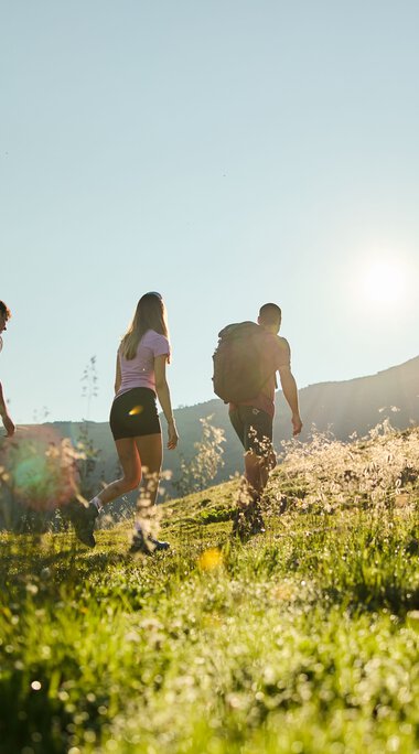 Drei Wanderer in Bewegung auf einem grünen Hang im Kleinwalsertal bei Sonnenaufgang, mit Berggipfeln im Hintergrund. | © Kleinwalsertal Tourismus eGen • Fotograf: Oliver Farys