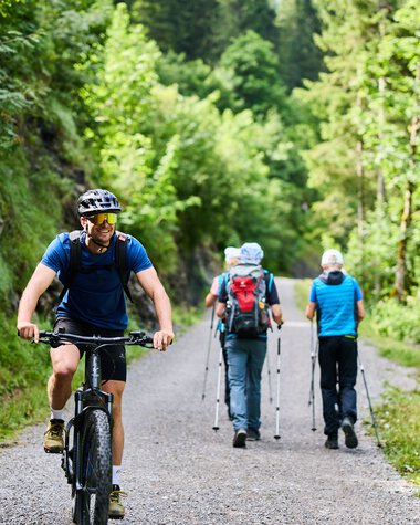 Four hikers with backpacks and trekking poles on a summer forest trail, one riding a bike. | © Kleinwalsertal Tourismus eGen • Photographer: Oliver Farys