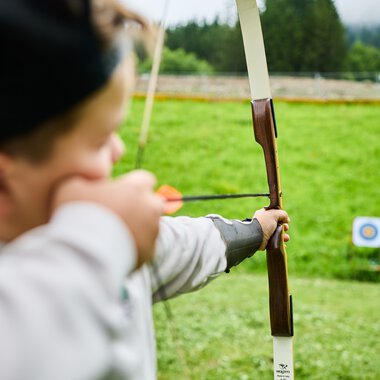 Ein Kind beim Bogenschießen in einer Wildnisschule im Kleinwalsertal, das Ziel im Hintergrund, die Natur ist grün und offen. | © Kleinwalsertal Tourismus eGen • Fotograf: Oliver Farys