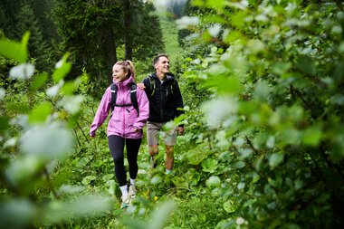 Ein Mann und eine Frau wandern zusammen durch einen grünen Wald im Sommer, lächeln und genießen die Natur. | © Kleinwalsertal Tourismus eGen • Fotograf: Oliver Farys