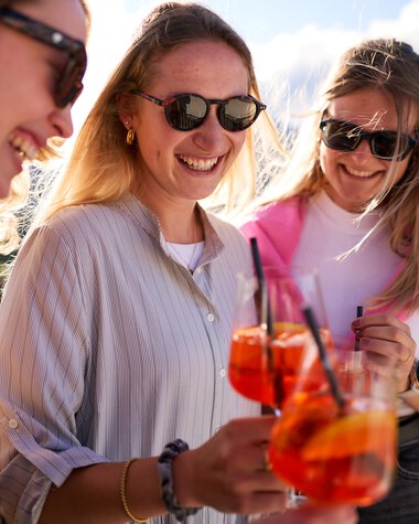Drei Frauen lächeln und stoßen mit Aperol Spritz auf einer Sommerlounge im Kleinwalsertal an. | © Kleinwalsertal Tourismus eGen • Fotograf: Oliver Farys