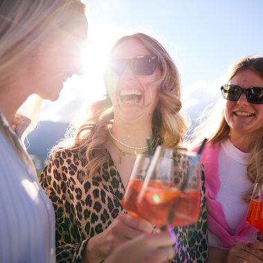 Drei lachende Frauen bei sommerlicher Lounge-Veranstaltung im Kleinwalsertal, trinken roséfarbene Getränke im Sonnenlicht. | © Kleinwalsertal Tourismus eGen • Fotograf: Oliver Farys