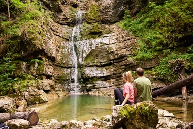 Zwei Menschen sitzen am Wasserfall im Kleinwalsertal, umgeben von grüner Natur, mit Blick auf den Wasserfall und das ruhige Wasser. | © Kleinwalsertal Tourismus eGen • Fotograf: Oliver Farys