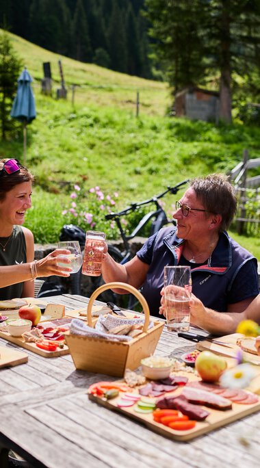 Vier Freunde sitzen an einem Holztisch im Grünen, genießen Getränke und Grillgut bei schönem Wetter in den Bergen, im Hintergrund Wiesen, Bäume und ein Haus. | © Kleinwalsertal Tourismus eGen • Fotograf: Oliver Farys