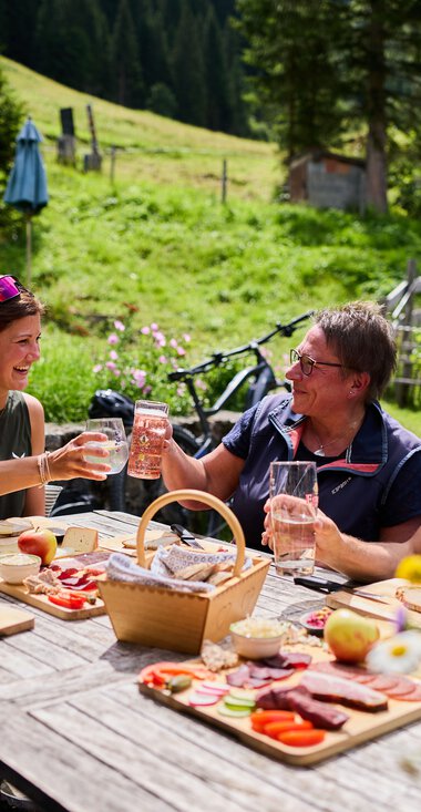 Vier Freunde sitzen an einem Holztisch im Grünen, genießen Getränke und Grillgut bei schönem Wetter in den Bergen, im Hintergrund Wiesen, Bäume und ein Haus. | © Kleinwalsertal Tourismus eGen • Fotograf: Oliver Farys