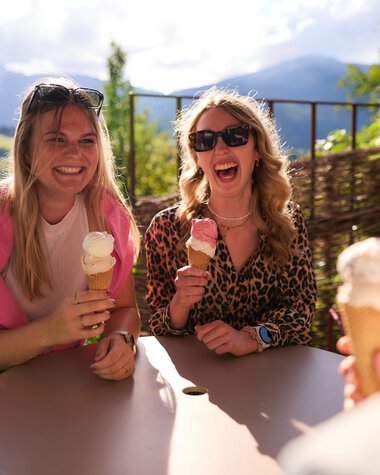 Drei Frauen sitzen im Sommer draußen am Tisch und essen Eis, lachen und genießen die Sonne mit schöner Bergkulisse im Hintergrund. | © Kleinwalsertal Tourismus eGen • Fotograf: Oliver Farys
