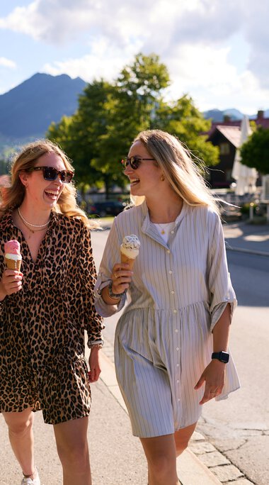 Drei junge Frauen gehen fröhlich im Sommer in Kleinwalsertal spazieren, jede hat ein Eis in der Hand, umgeben von grünen Bäumen und Bergen im Hintergrund. | © Kleinwalsertal Tourismus eGen • Fotograf: Oliver Farys