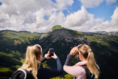 Zwei Frauen fotografieren eine beeindruckende Bergkulisse im Kleinwalsertal, umgeben von Wäldern und bewölktem Himmel. | © Kleinwalsertal Tourismus eGen • Fotograf: Oliver Farys