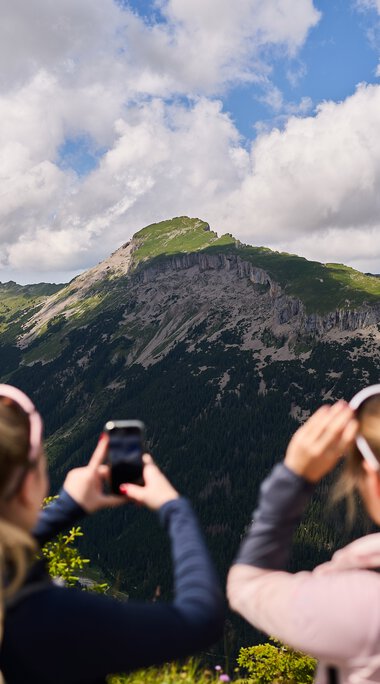 Zwei Frauen fotografieren eine beeindruckende Bergkulisse im Kleinwalsertal, umgeben von Wäldern und bewölktem Himmel. | © Kleinwalsertal Tourismus eGen • Fotograf: Oliver Farys