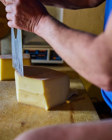 Nahaufnahme eines Mannes, der einen Block Käse mit einem großen Messer auf einem Holztisch schneidet, im Hintergrund eine Käseform. | © Kleinwalsertal Tourismus eGen • Fotograf: Oliver Farys