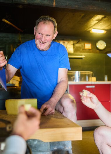 Mann in blauer T-Shirt hält einen Zettel und unterhält sich mit zwei anderen Personen in einem gemütlichen Raum. | © Kleinwalsertal Tourismus eGen • Fotograf: Oliver Farys