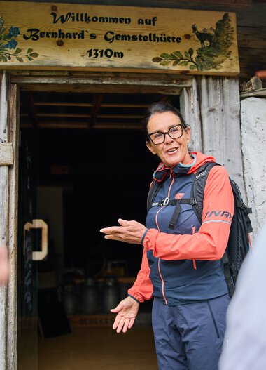 Frau begrüßt eine Gruppe von Wanderern vor der Käse- und Gemststätte im Kleinwalsertal, umgeben von Berglandschaft. | © Kleinwalsertal Tourismus eGen • Fotograf: Oliver Farys