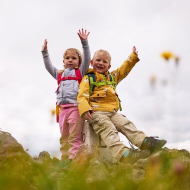 Two smiling children hiking in a mountainous landscape, reaching a summit with clouds in the background. | © Kleinwalsertal Tourismus eGen • Photographer: Oliver Farys
