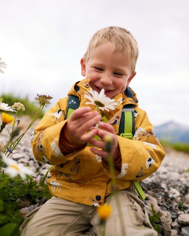 Junge beim Wandern im Kleinwalsertal, lachend mit einer Blume in der Hand, umgeben von Blumen und Natur. | © Kleinwalsertal Tourismus eGen • Fotograf: Oliver Farys