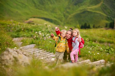 Familie mit zwei Kindern wandert auf einem Holzsteg in einer grünen Berglandschaft im Kleinwalsertal. | © Kleinwalsertal Tourismus eGen • Fotograf: Oliver Farys