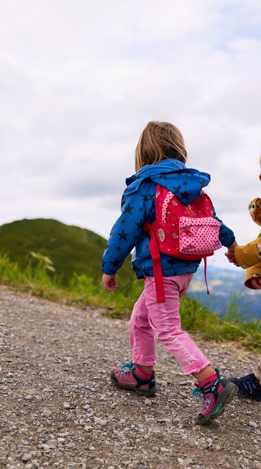 Zwei Kinder wandern auf einem Bergpfad, eines schaut nach hinten und lächelt, getragen von bunten Rucksäcken, im Sommer in den Bergen. | © Kleinwalsertal Tourismus eGen • Fotograf: Oliver Farys