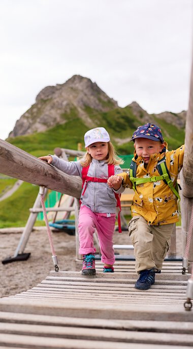 Zwei Kinder schaukeln auf einer Hängebrücke im Kleinwalsertal, umgeben von Bergen. | © Kleinwalsertal Tourismus eGen • Fotograf: Oliver Farys